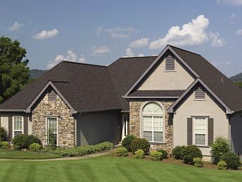 House with stone and stucco exterior, dark roof, and green lawn under a blue sky.