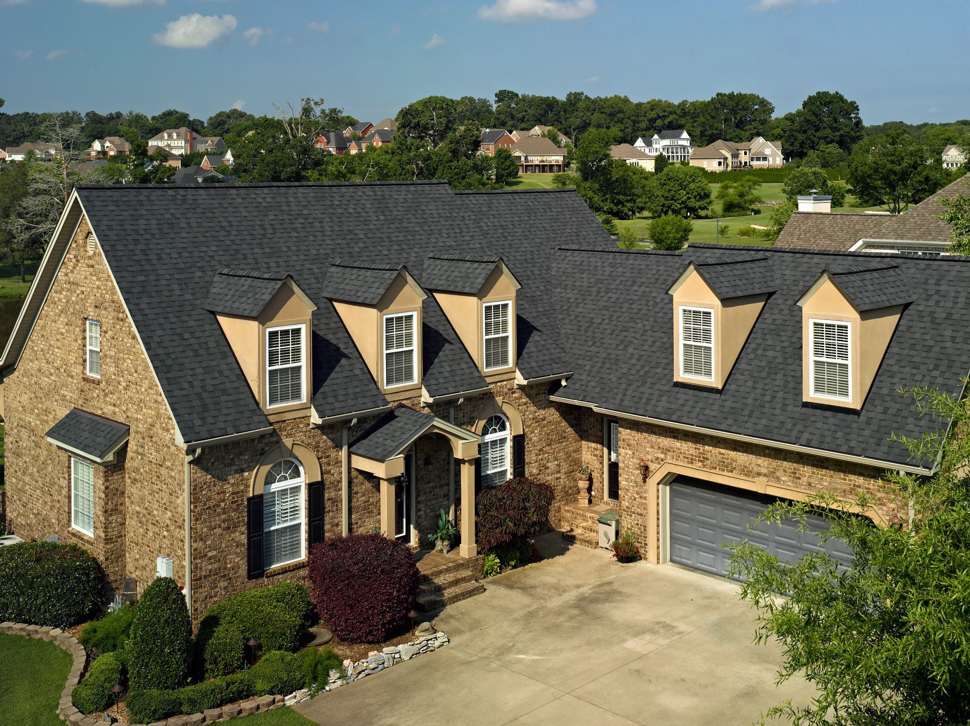 Brick house with dark gray roof and dormers, surrounded by greenery and other houses in the background.