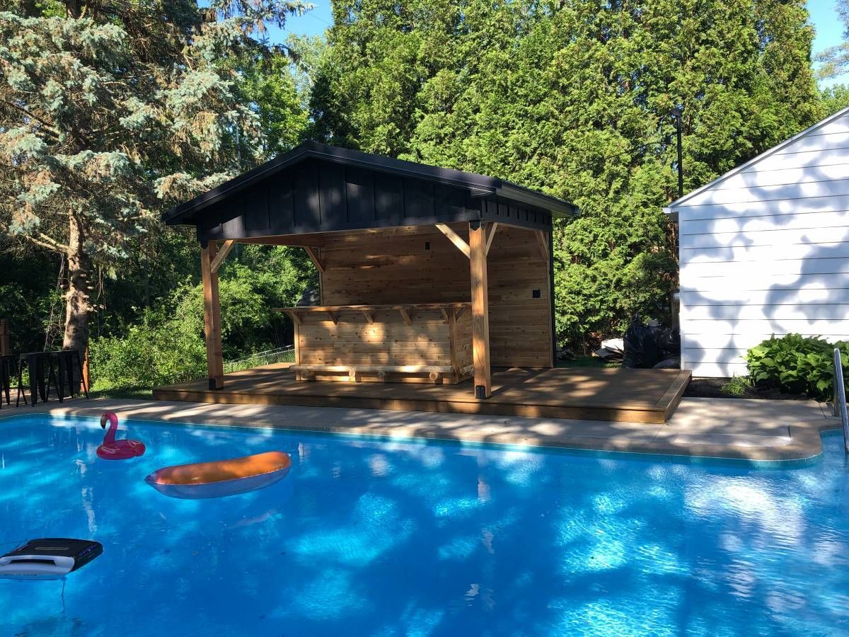 A wooden outdoor bar under a black-roofed pavilion beside a bright blue swimming pool with floating pool toys.