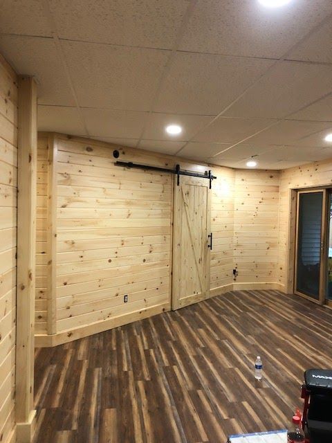 A room with knotty pine walls, dark wood-look flooring, and a sliding barn-style door mounted on black hardware.