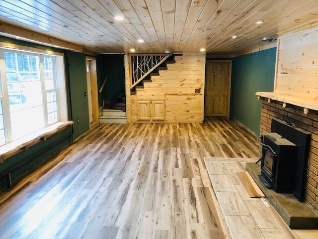 A rustic room with wood-paneled ceiling, light wood floors, a brick fireplace with a stove, and a window seat.