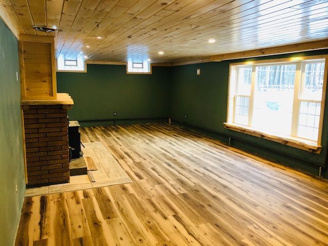 A renovated basement room with wood-paneled ceilings, green walls, light wood flooring, a brick fireplace, & a large window.