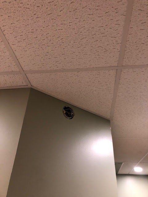 A small, circular vent or port is located on a beige wall just below a drop ceiling in an indoor space.