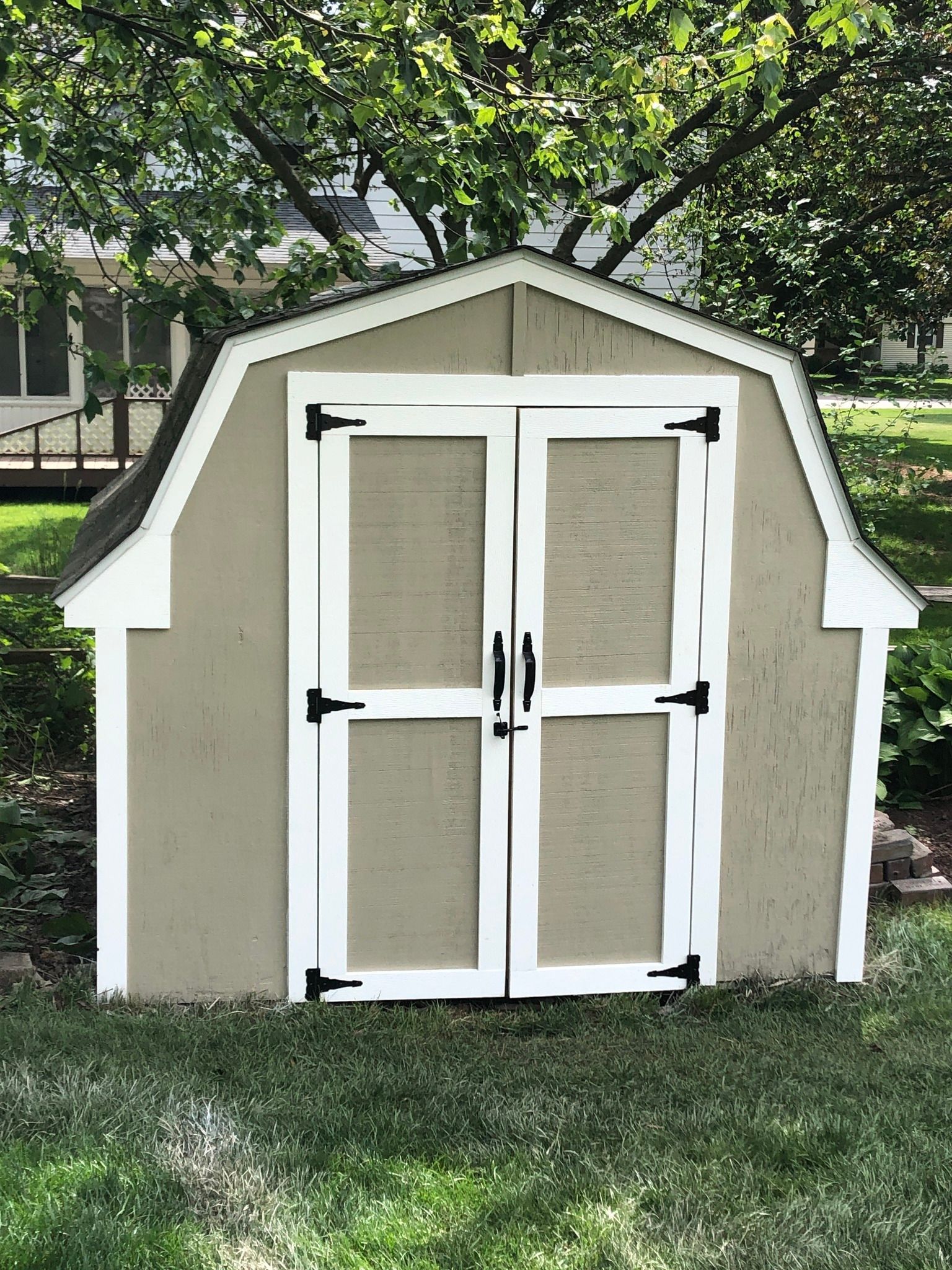A beige wooden storage shed with white trim and double doors, situated on a grassy lawn under trees.