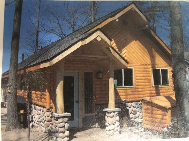 A rustic log cabin with a stone-based porch, shingled roof, and large windows, surrounded by trees on a sunny day.
