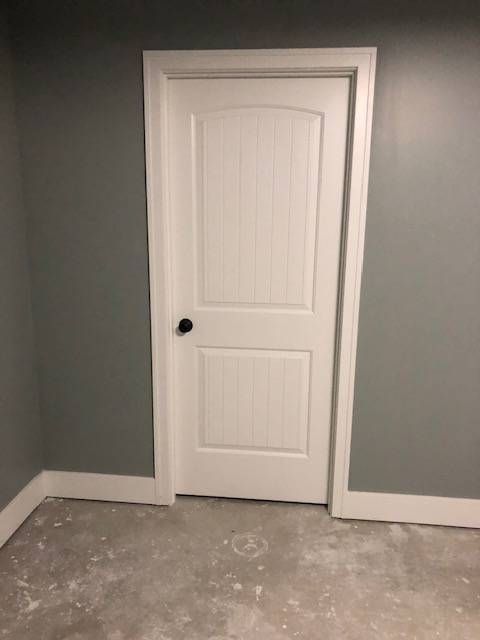 A white paneled door with a black knob set in a gray wall above unfinished concrete flooring.