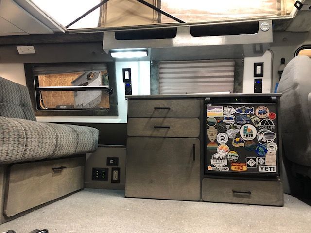 Interior of a camper featuring a fabric bench seat next to a storage cabinet and a refrigerator covered in stickers.