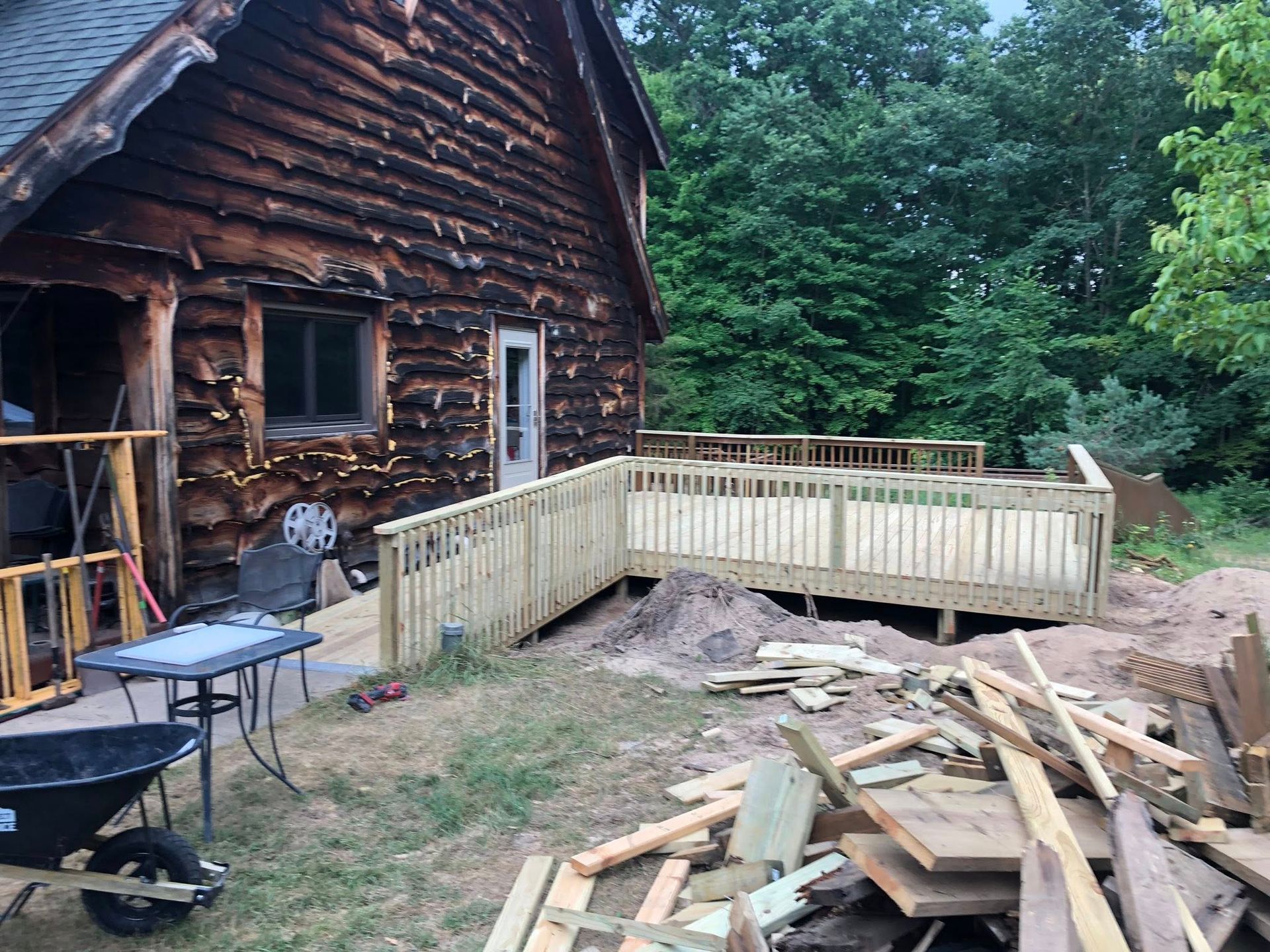A wooden deck with railings under construction next to a rustic log cabin, with building materials scattered on the ground.