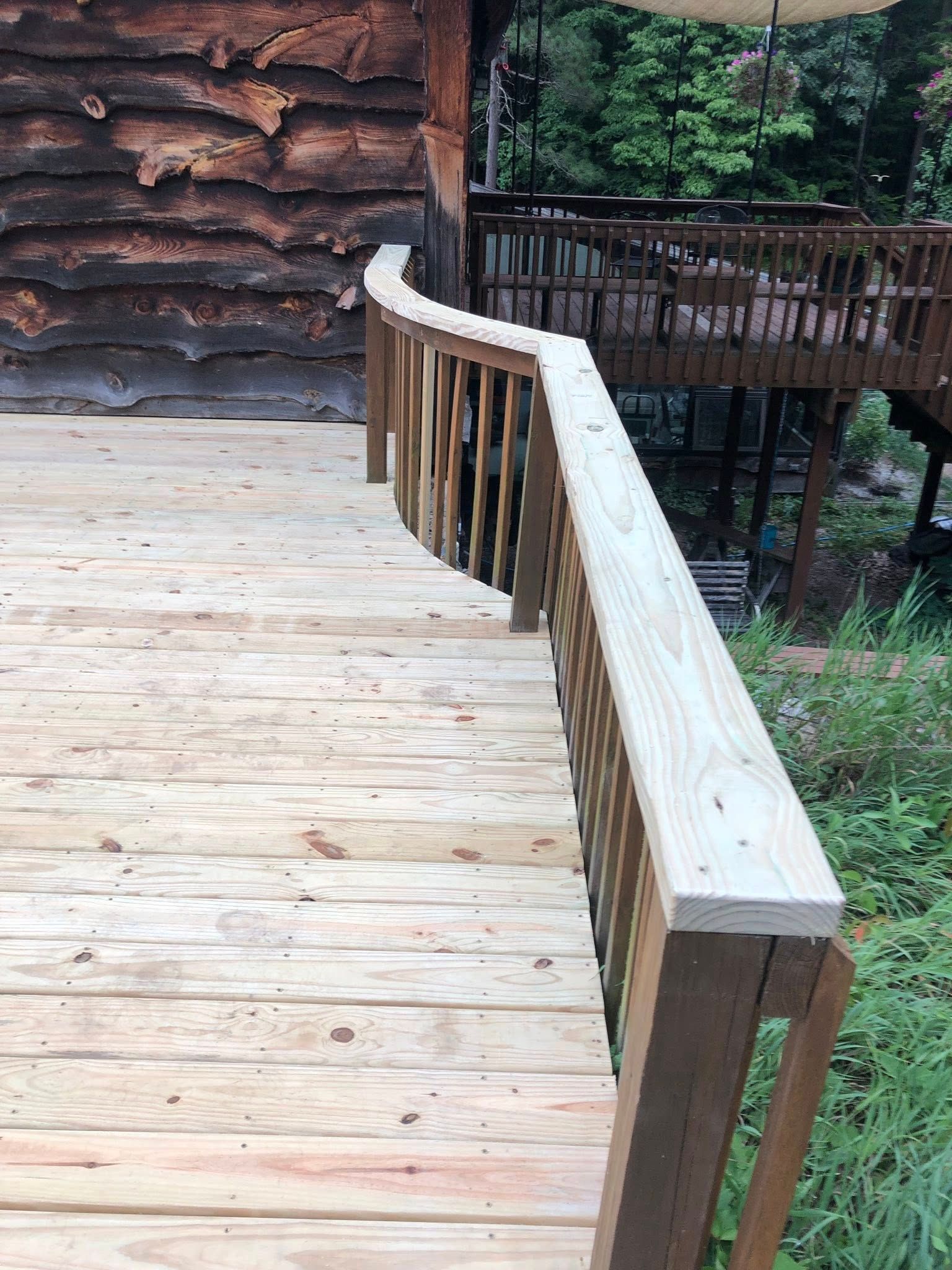 A light-colored wooden deck curves around a rustic cabin, featuring a matching railing and a view of trees.