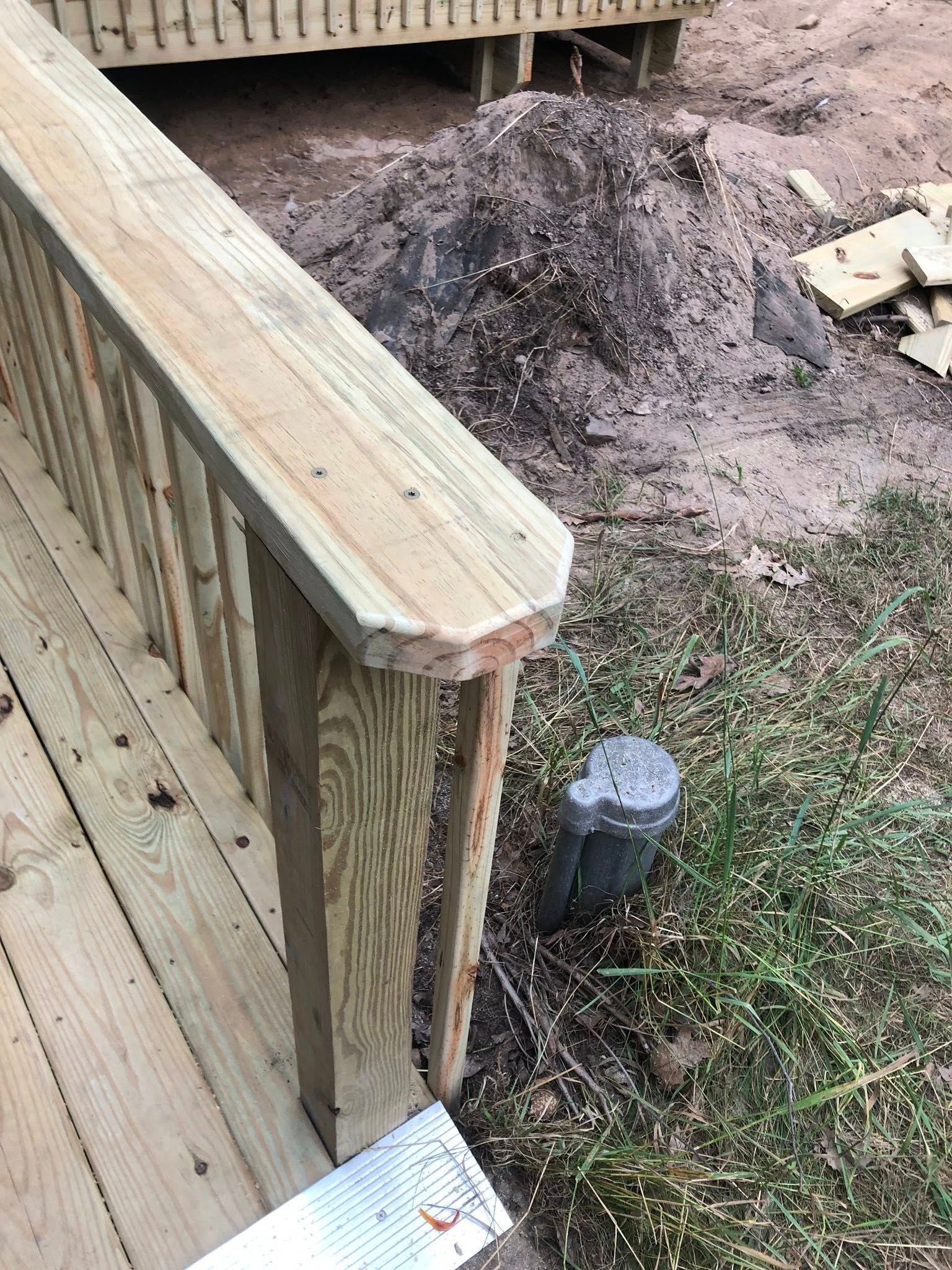 A close-up of a wooden deck railing end cap overlooking a dirt construction site with a utility pipe in the grass below.