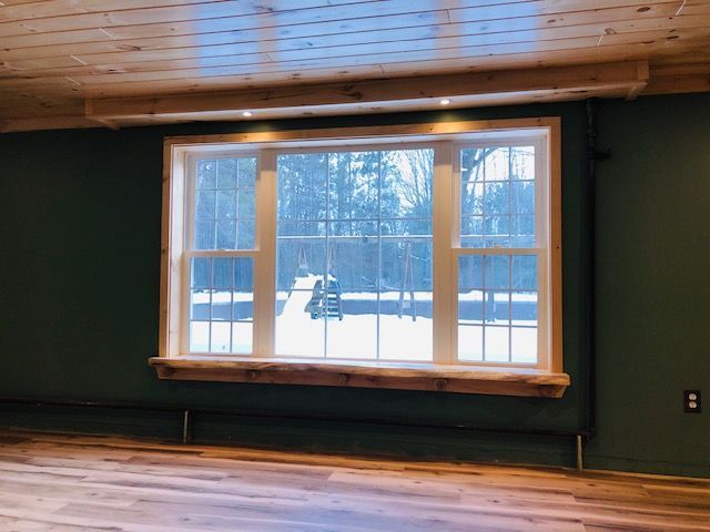 A window with a natural wood ledge and frame set in a dark green wall above wood flooring, overlooking a snowy yard.