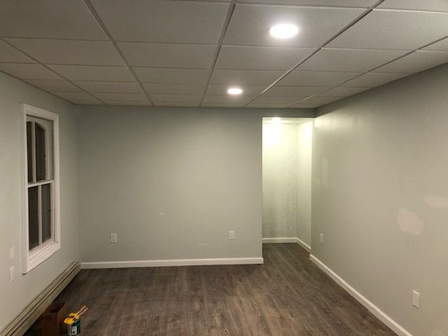 A room with light green walls, a white drop ceiling with recessed lights, and wood-look flooring, with a window on the left.