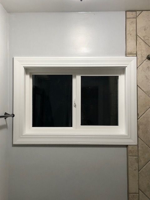 A white rectangular horizontal window with two panes centered on a light gray wall next to a beige tiled corner.