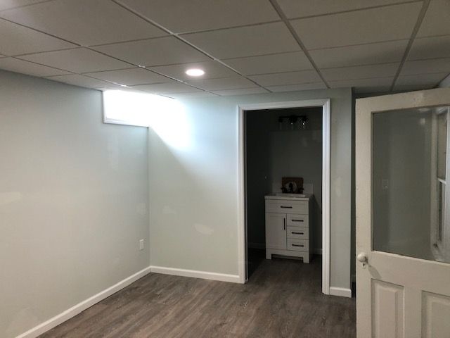 A basement room with light gray walls, wood-look flooring, a drop ceiling, a window, and a doorway leading to a vanity.