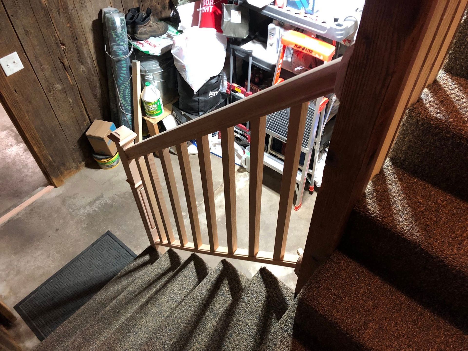 A wooden baby gate installed at the top of a carpeted staircase leading to a cluttered storage area.
