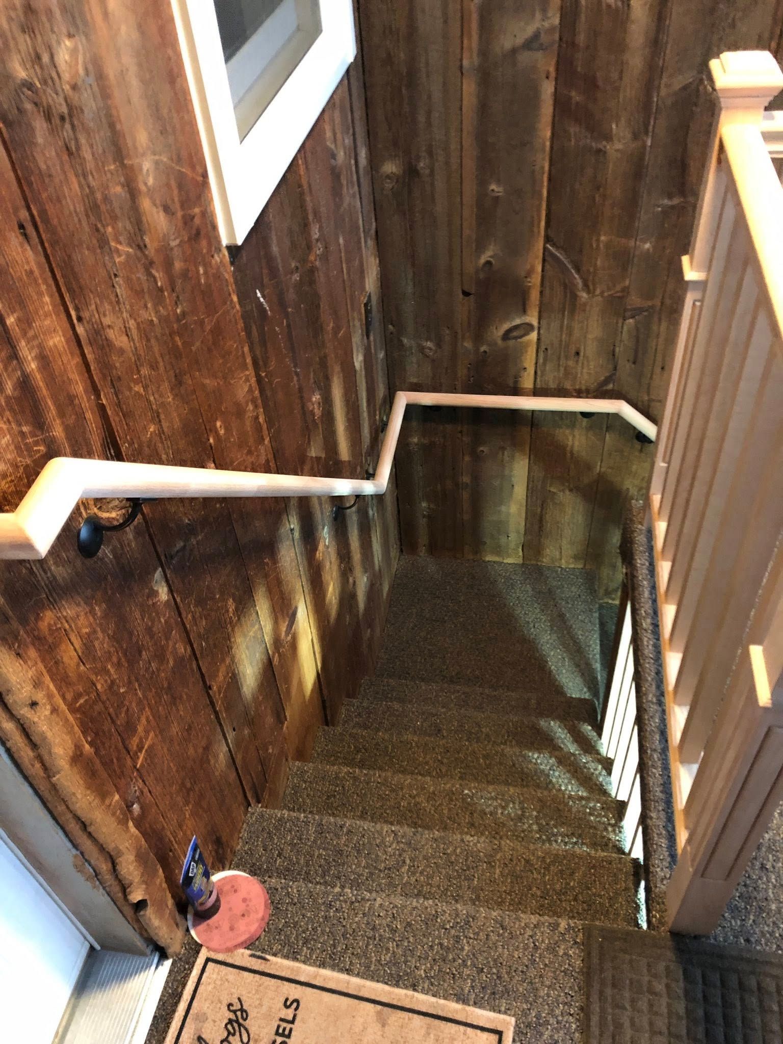 A narrow staircase with rustic wood walls, carpeted steps, and a white handrail that follows the turn of the stairs.