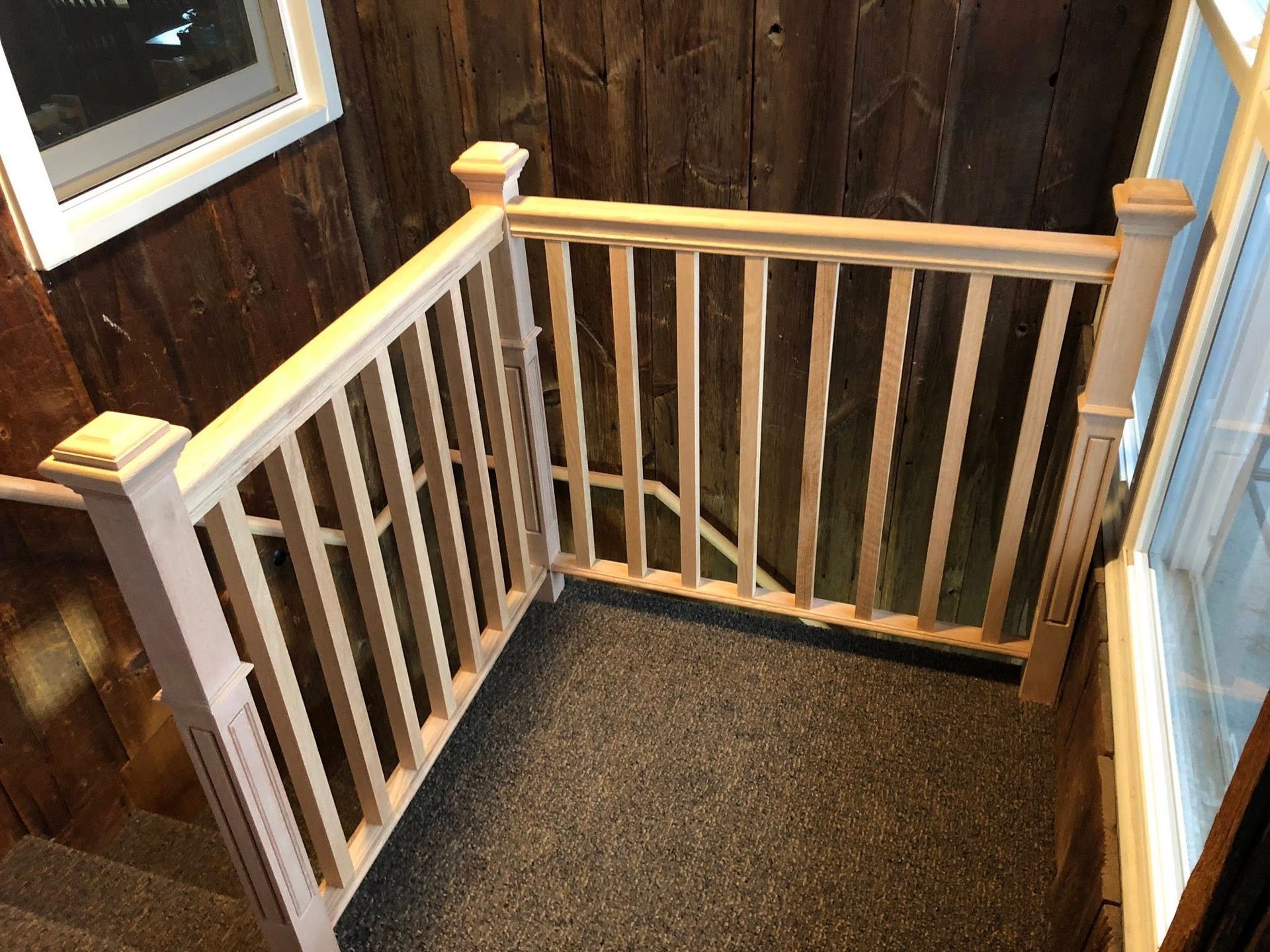 A light-colored wooden railing with vertical balusters and square posts installed at the top of carpeted stairs.
