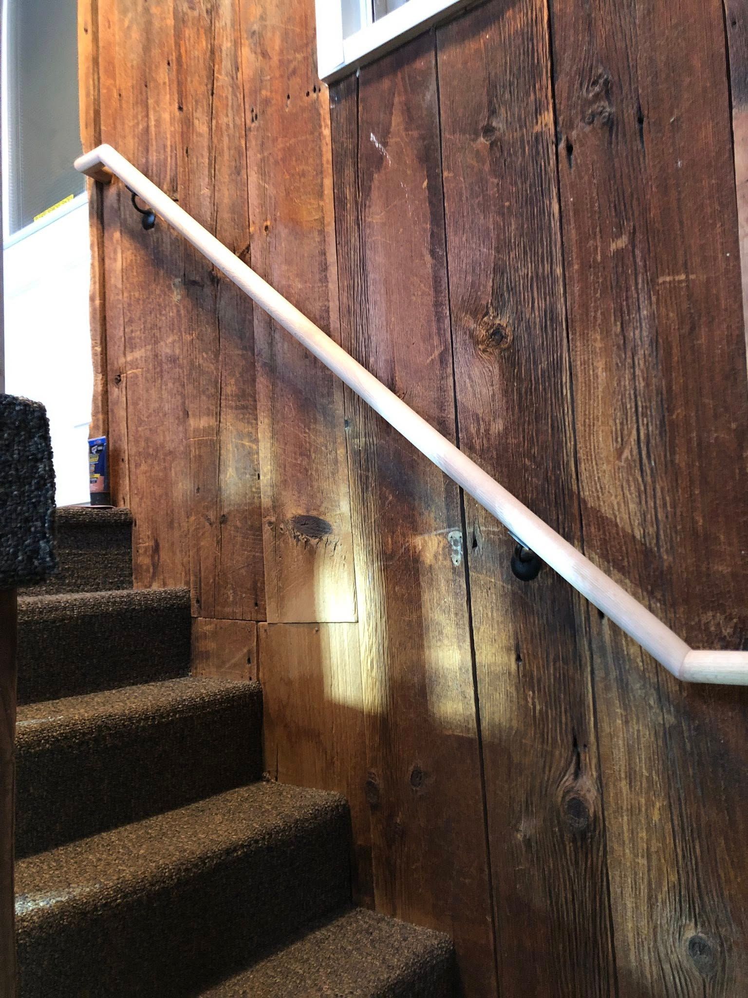 A white handrail runs diagonally along a wall made of weathered, brown wooden planks next to a carpeted staircase.