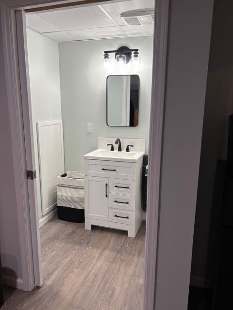 A small, modern bathroom with light green walls, wood-look flooring, a white vanity, and a black-framed mirror.