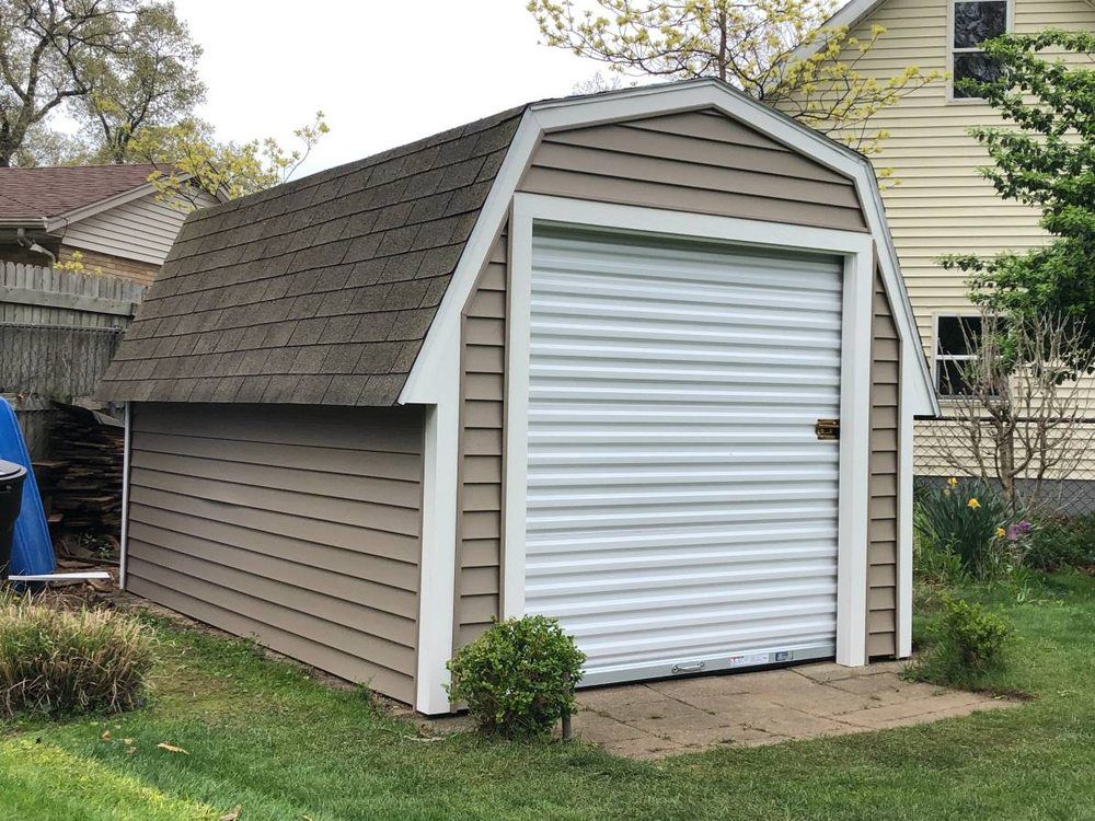 A tan, shed-style structure with a white roll-up door and dark shingles, situated on a grassy lawn.