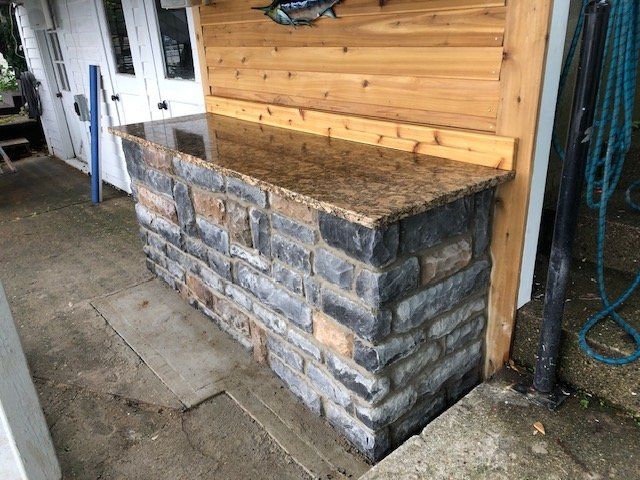 Outdoor stone bar with a polished brown granite countertop, positioned against a light wood panel wall.