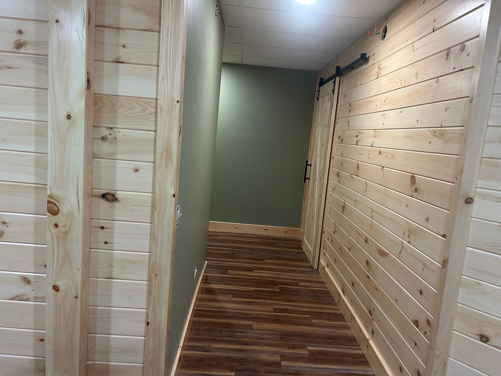 A narrow hallway with light wood paneling, a dark wood-look floor, an olive-green accent wall, and a sliding barn door.