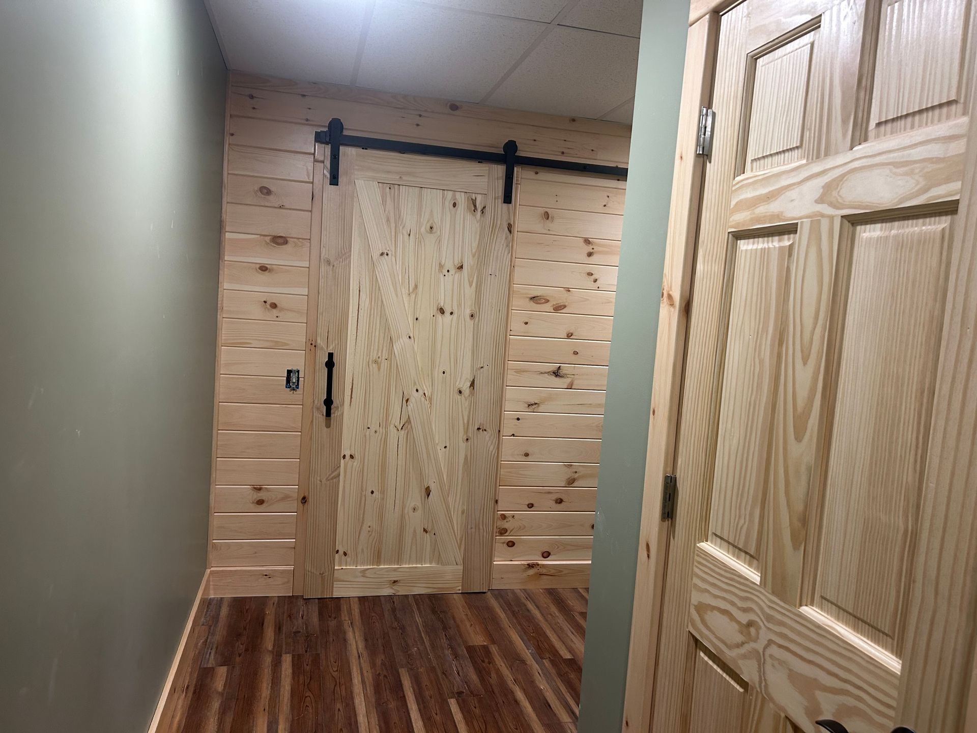 A hallway with a wooden barn door on a black rail, wood-paneled walls, and a matching six-panel door in the foreground.