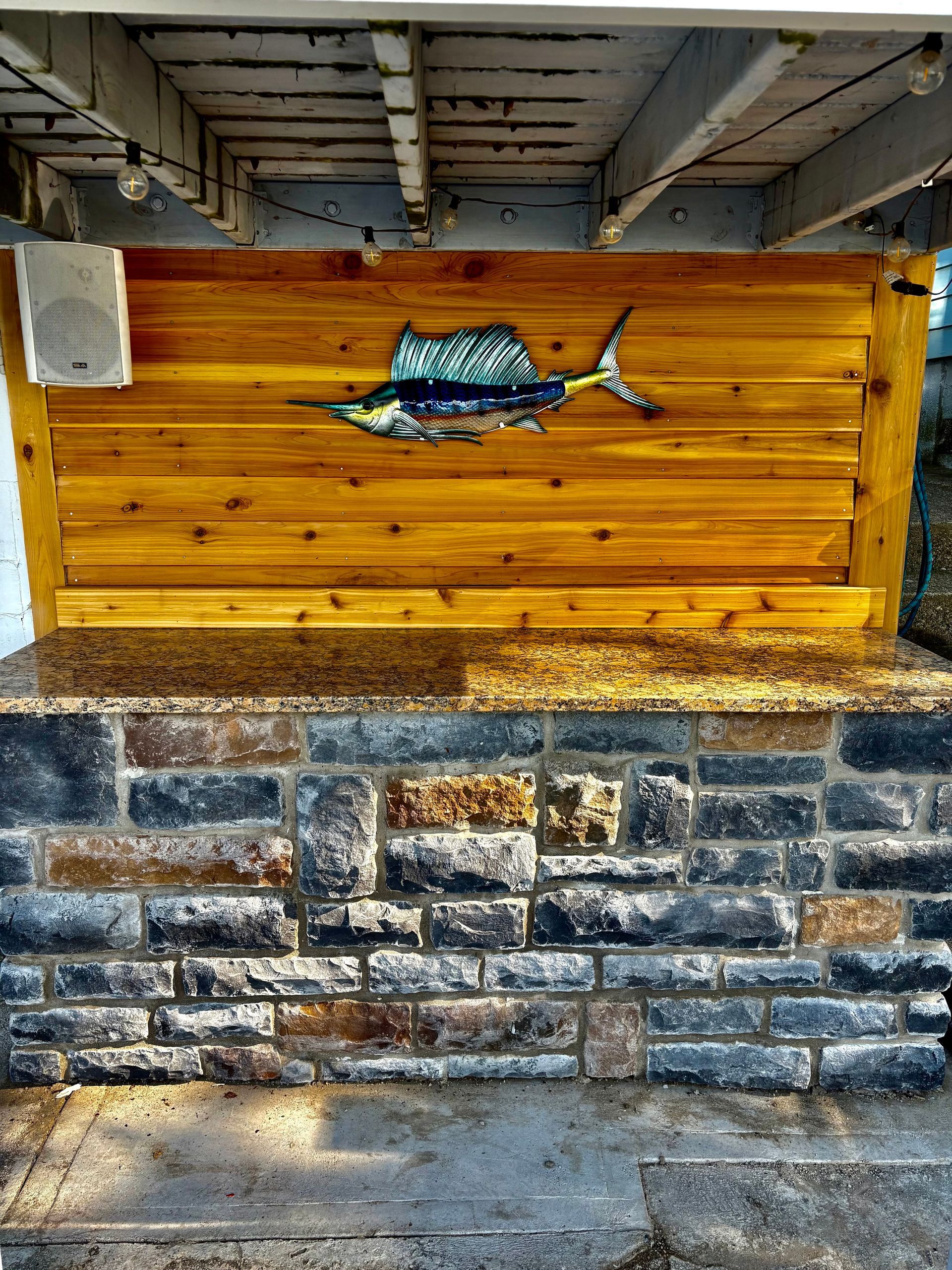 An outdoor bar with a stone base, granite countertop, and a wood-paneled wall featuring a sailfish wall hanging.