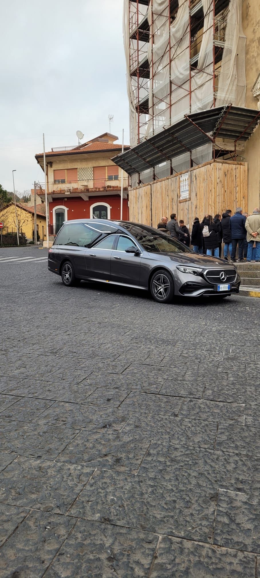 Carro funebre grigio su una strada acciottolata, edificio con impalcature sullo sfondo.