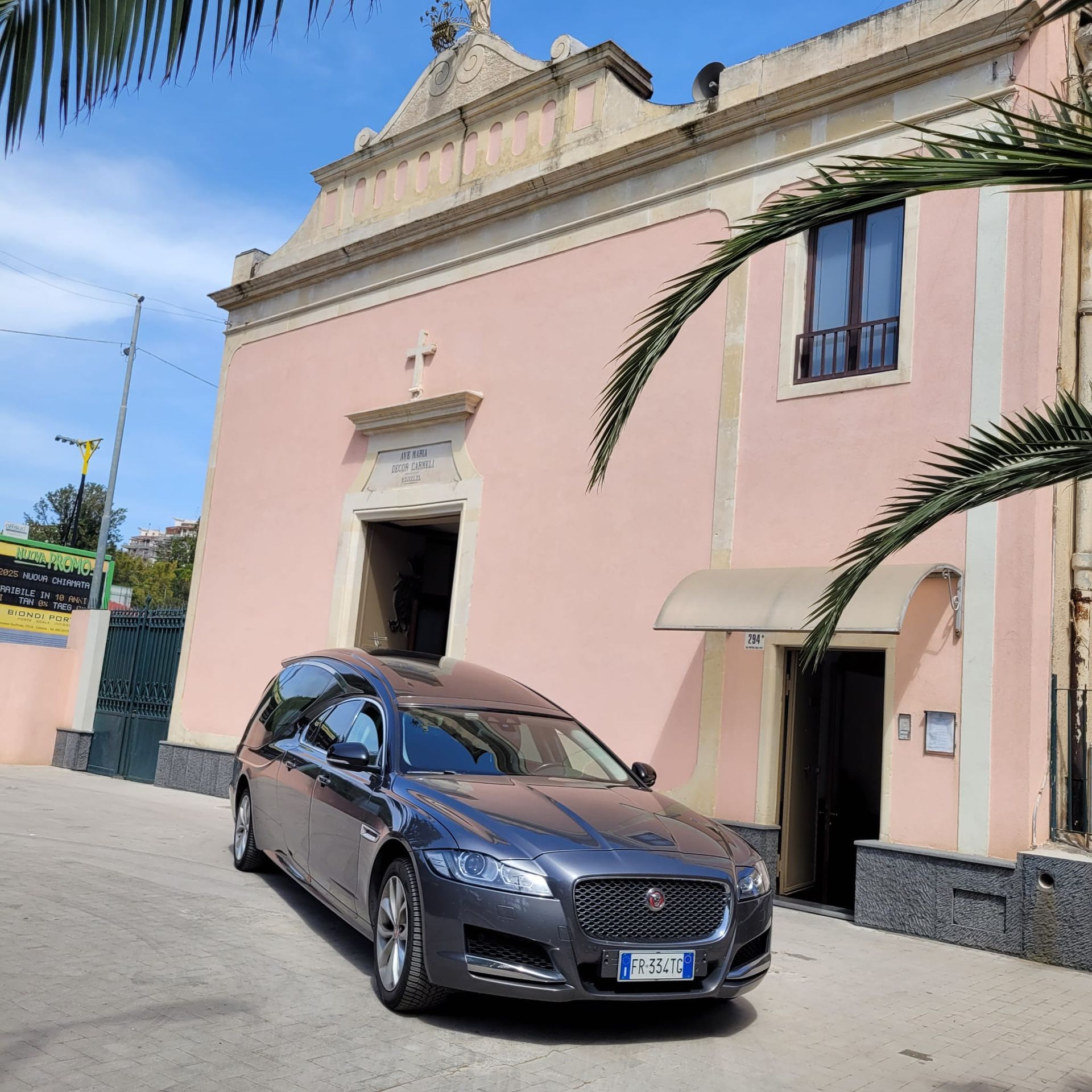 Carro funebre parcheggiato davanti a un edificio rosa con un ingresso ad arco e una croce in cima. Giornata di sole, palme.
