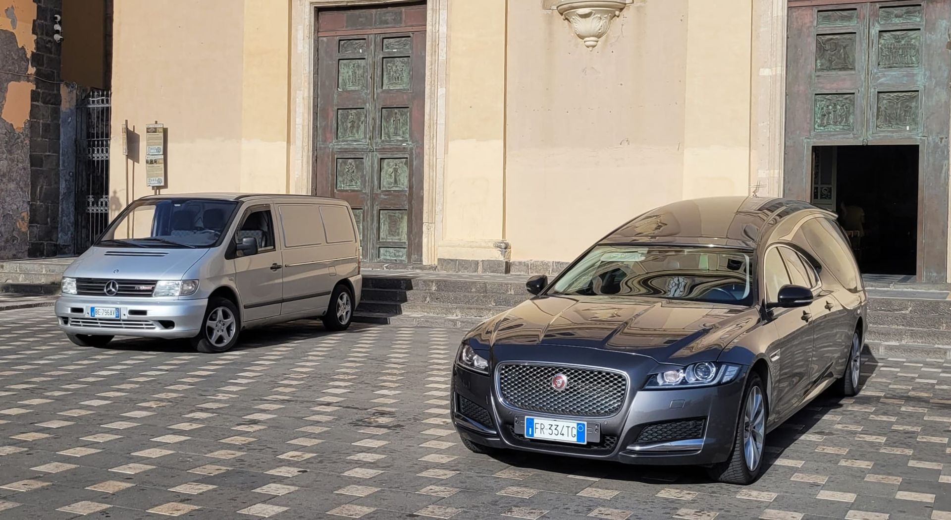Un furgone argentato e un carro funebre grigio scuro parcheggiati davanti a un edificio con la facciata in pietra.