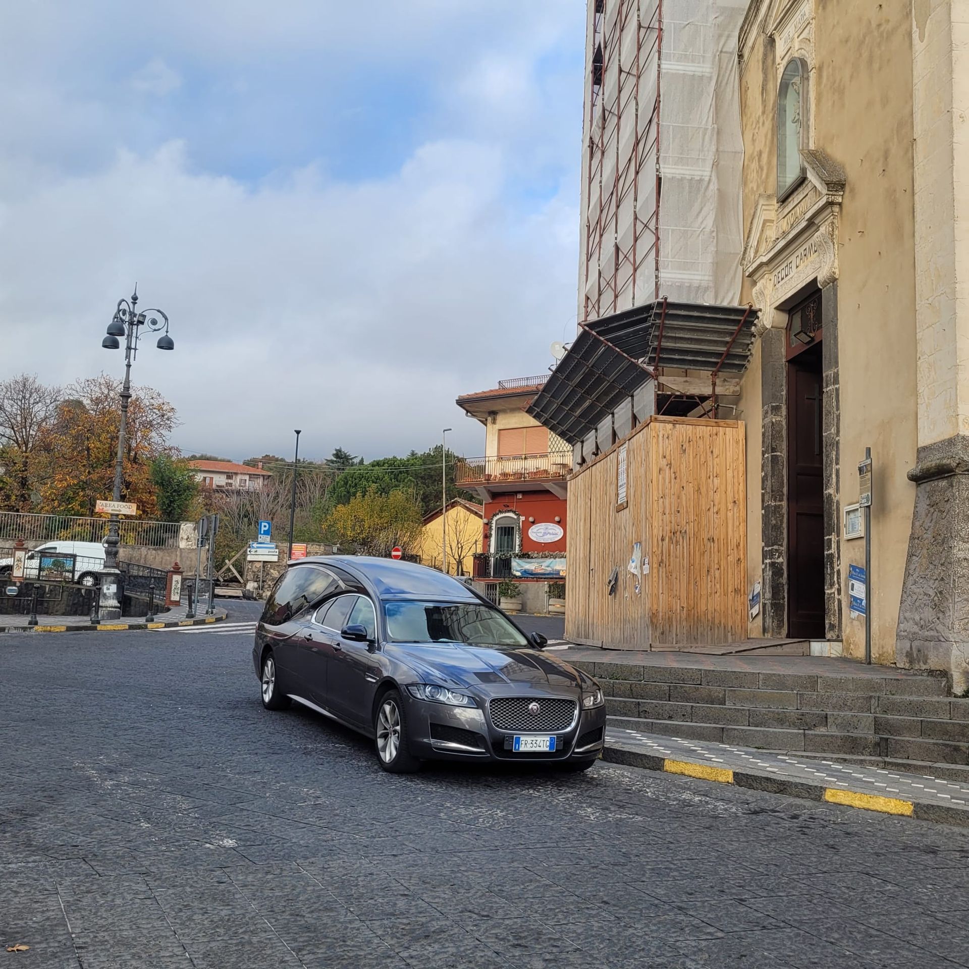 Un carro funebre parcheggiato su una strada acciottolata di fronte a una chiesa. Cielo nuvoloso.