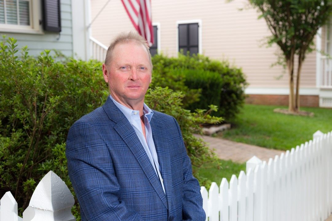 A man in a suit is standing next to a white picket fence in front of a house.