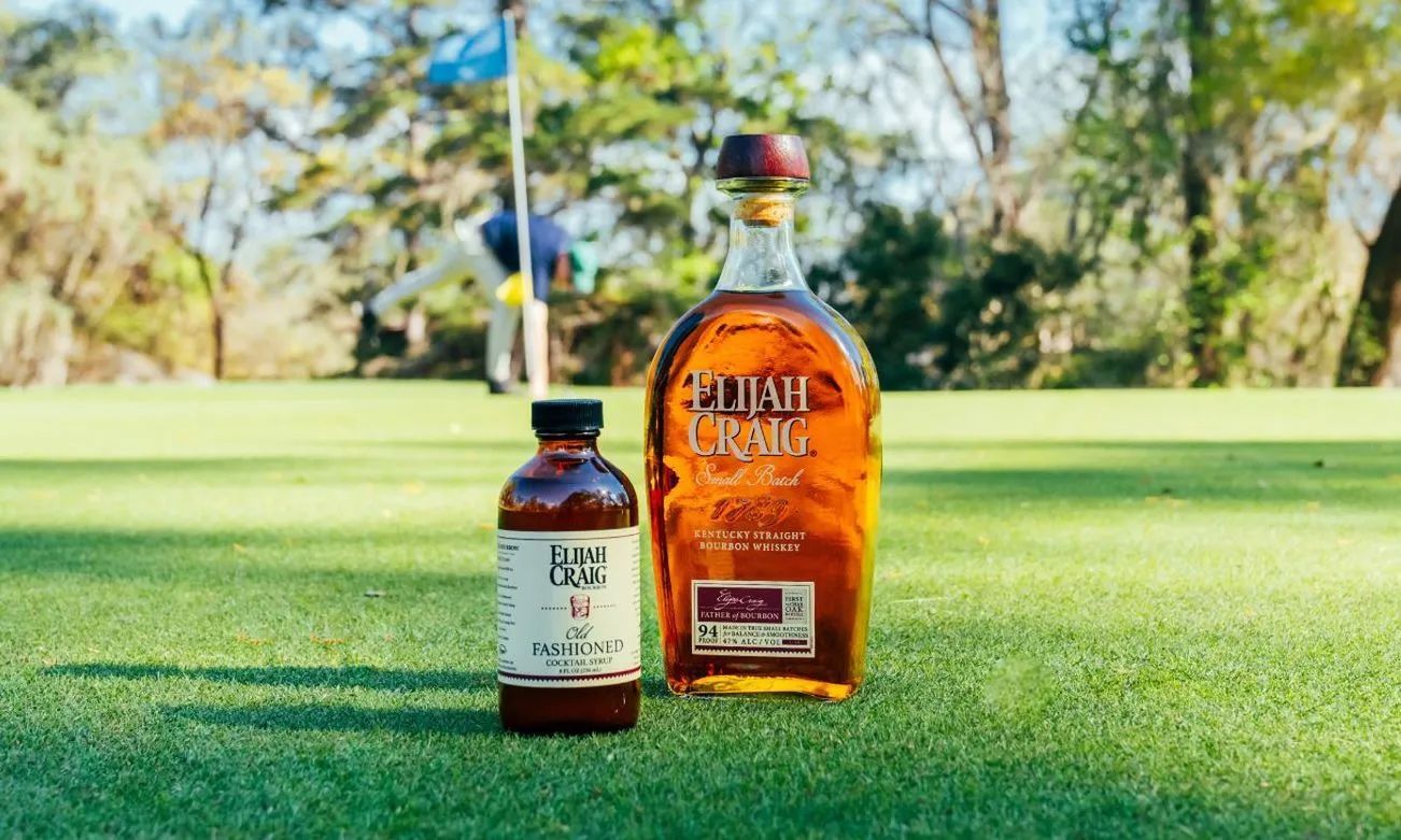 A bottle of whiskey and a bottle of bbq sauce are on a golf course.