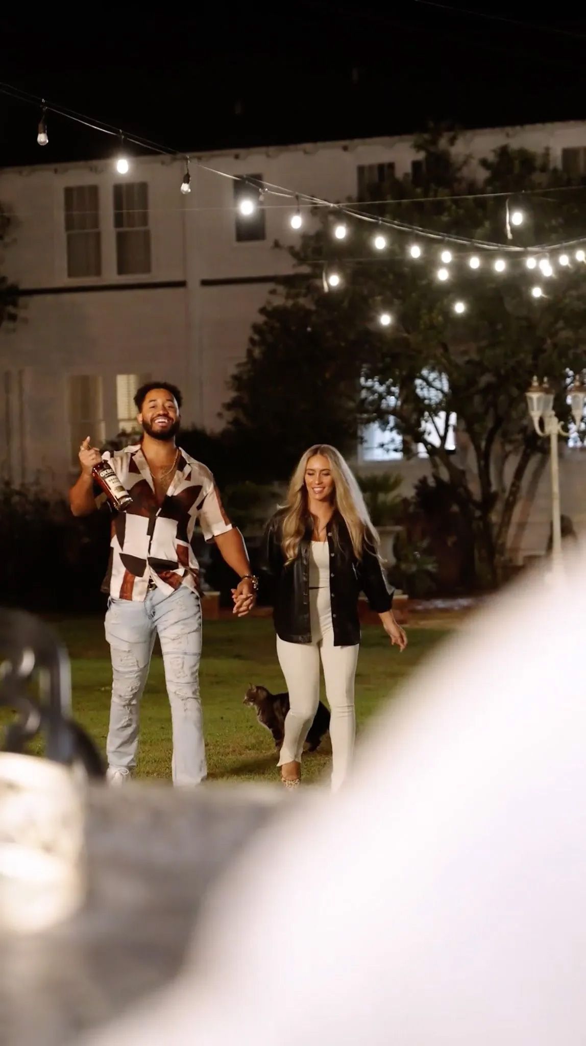 A man and a woman are holding hands and walking in front of a building at night.