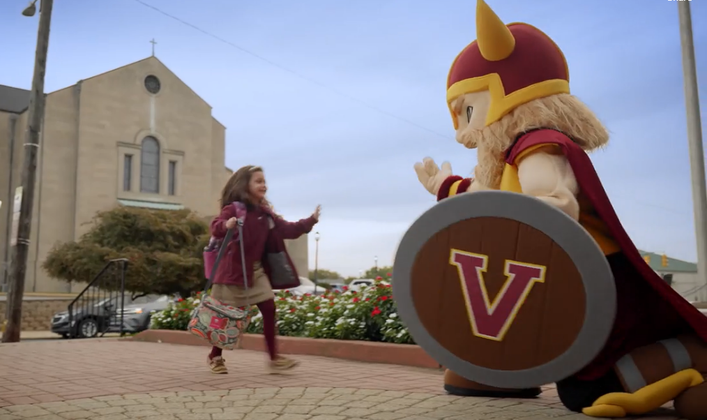 A girl is standing next to a viking mascot holding a shield with the letter v on it.