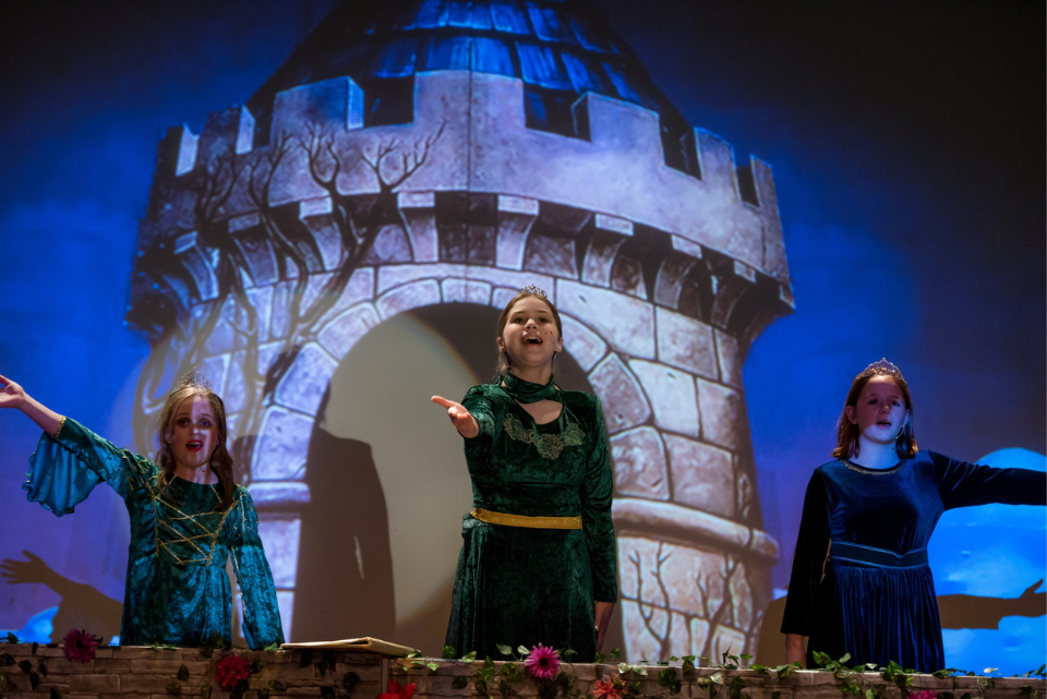 Three girls are standing on a stage in front of a castle.