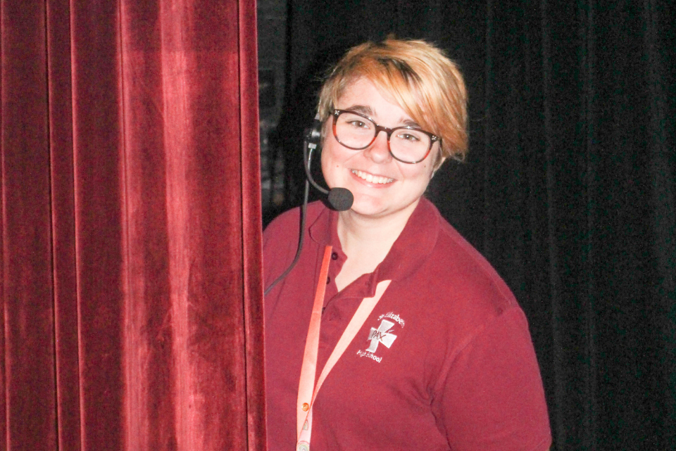 A woman wearing glasses and a headset stands in front of a red curtain