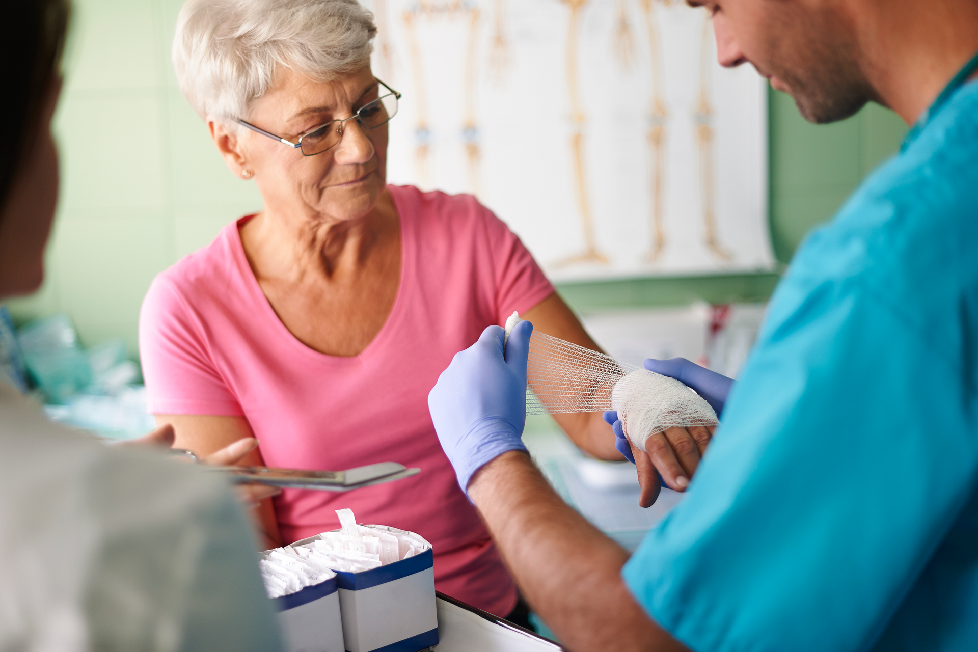 A male doctor is bandaging a senior woman on her hand with a nurse looking on.