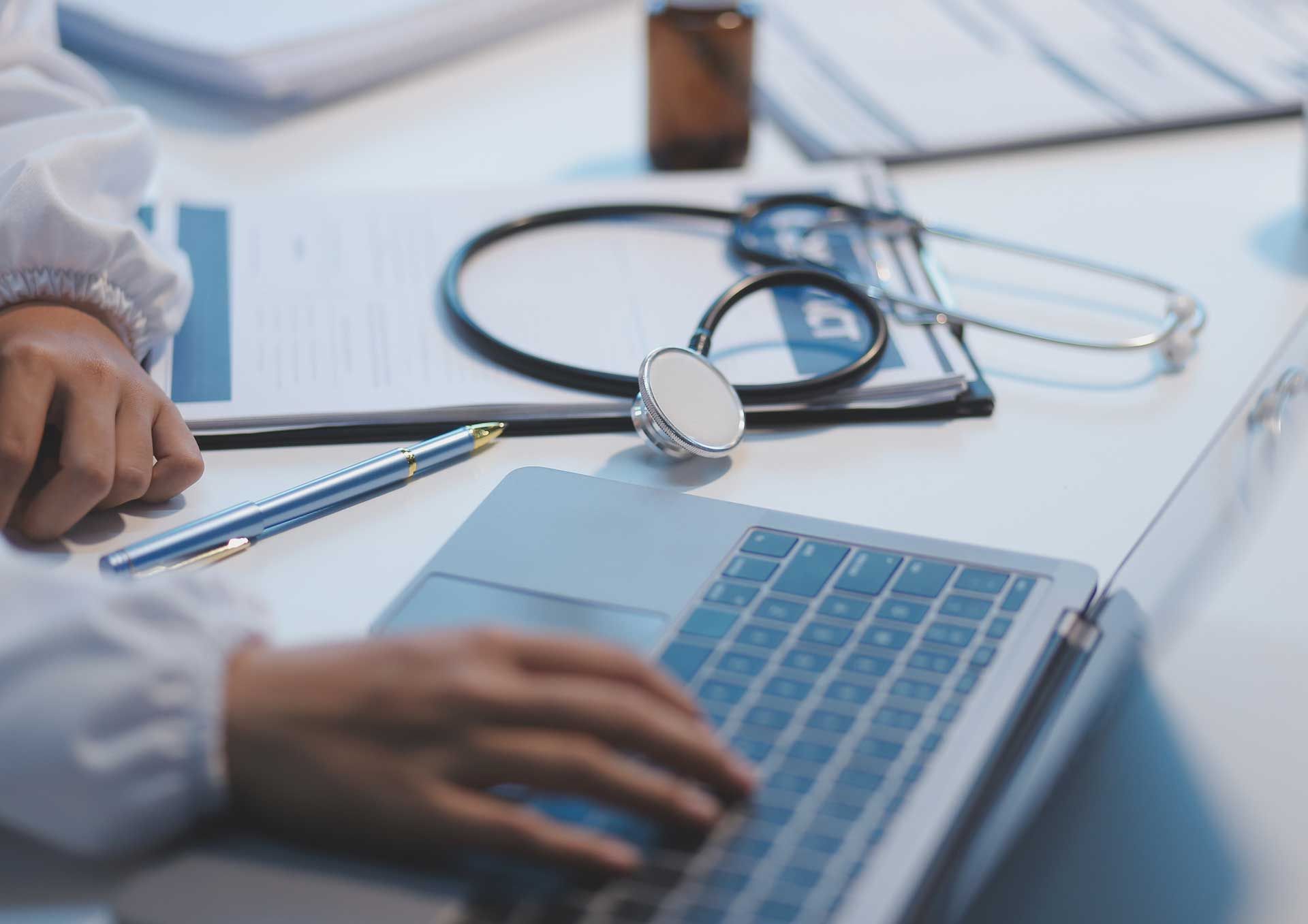 Doctor typing on laptop beside stethoscope and documents to help patients find medical clinic.