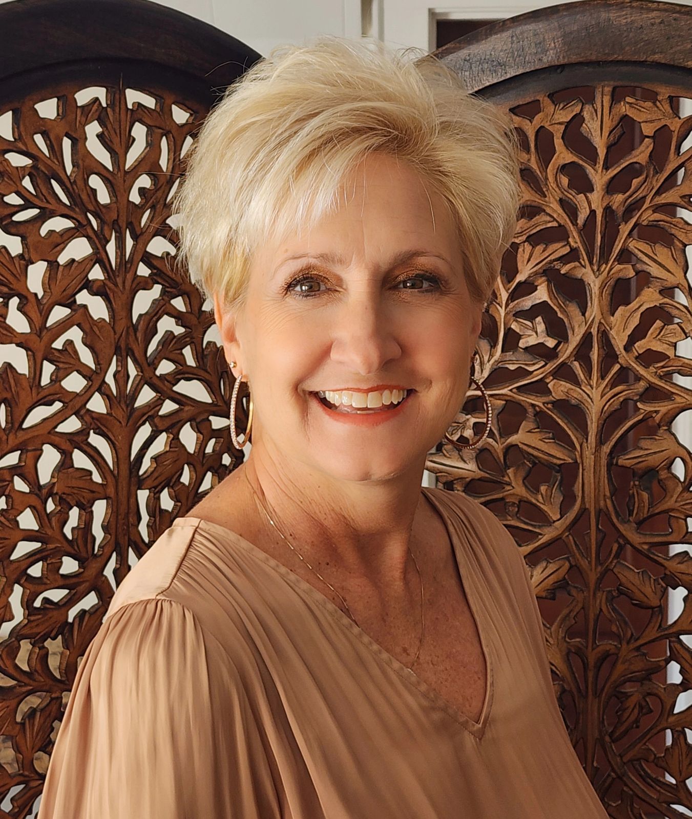 A woman in a brown shirt is smiling in front of a wooden screen.