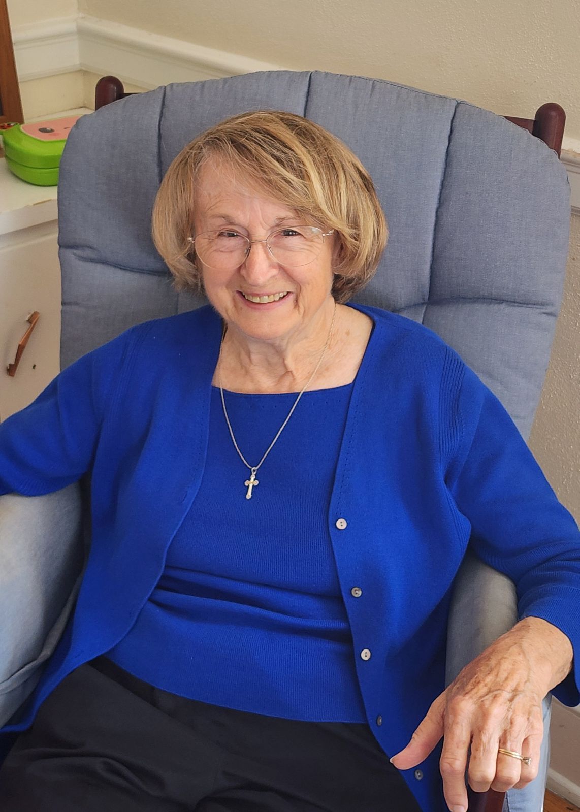 An elderly woman is sitting in a chair wearing a blue sweater and a cross necklace.