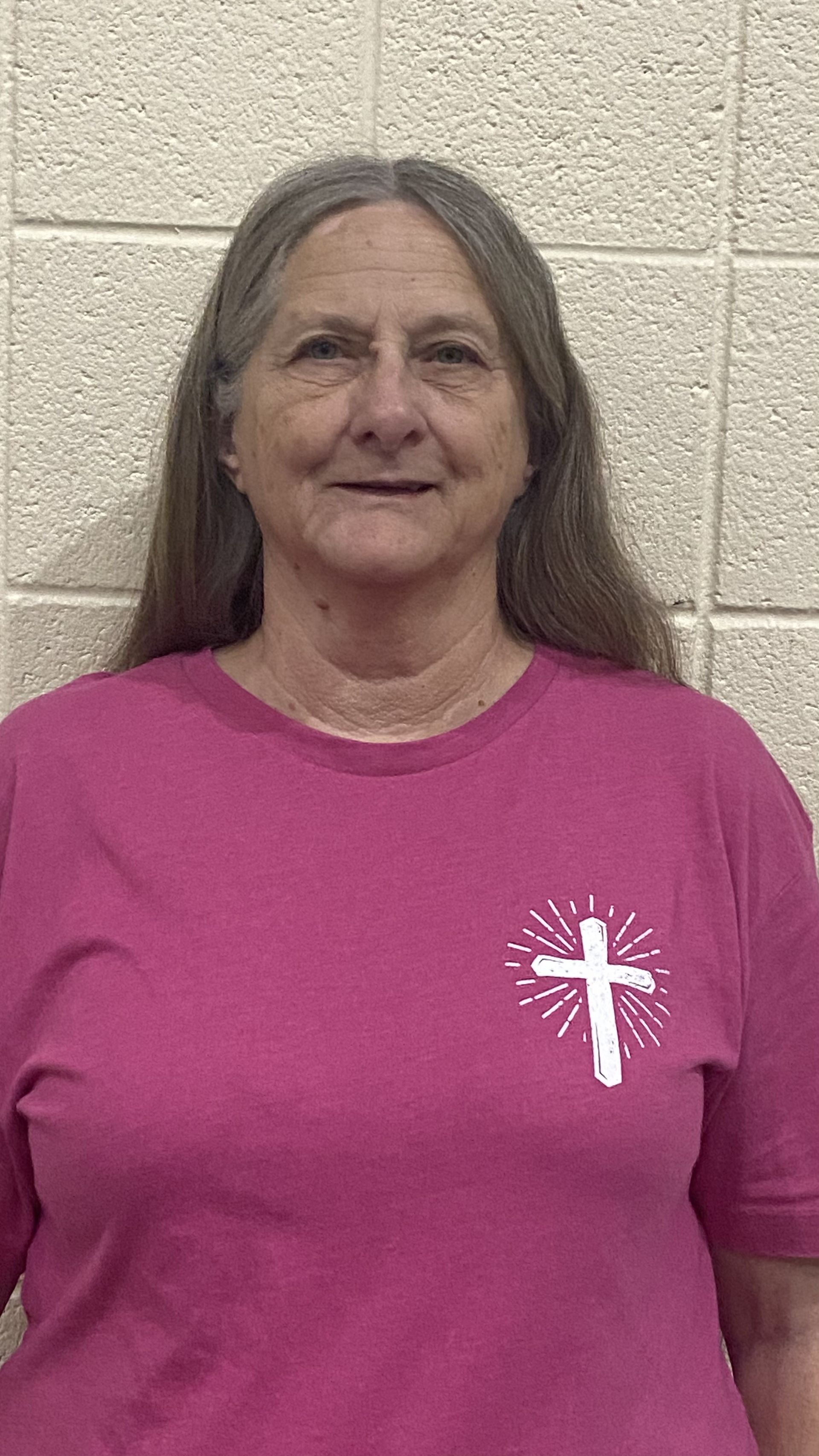 A woman wearing a pink t-shirt with a cross on it is standing in front of a brick wall.