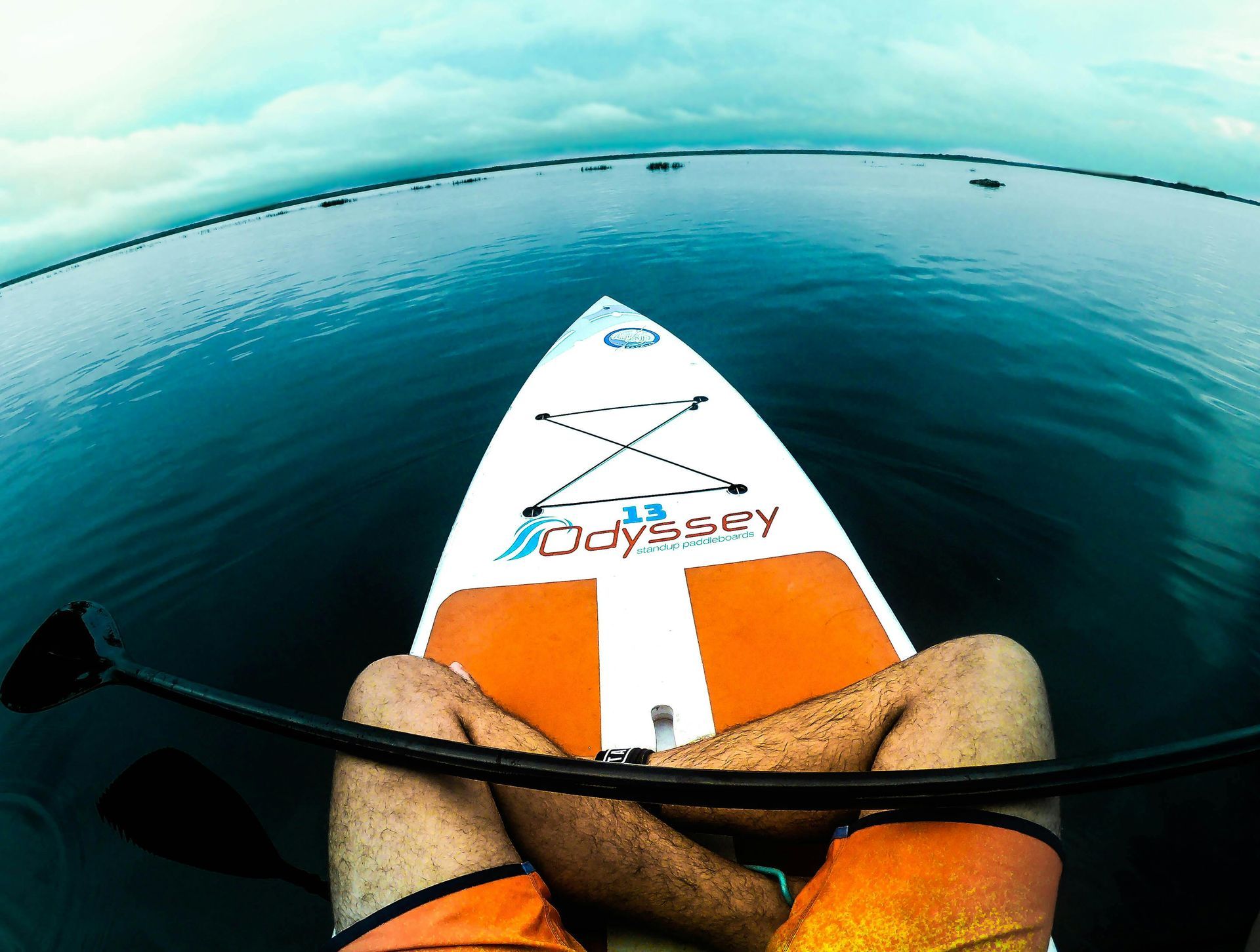 Paddleboard view over calm blue water, with orange shorts and a paddle in the foreground.