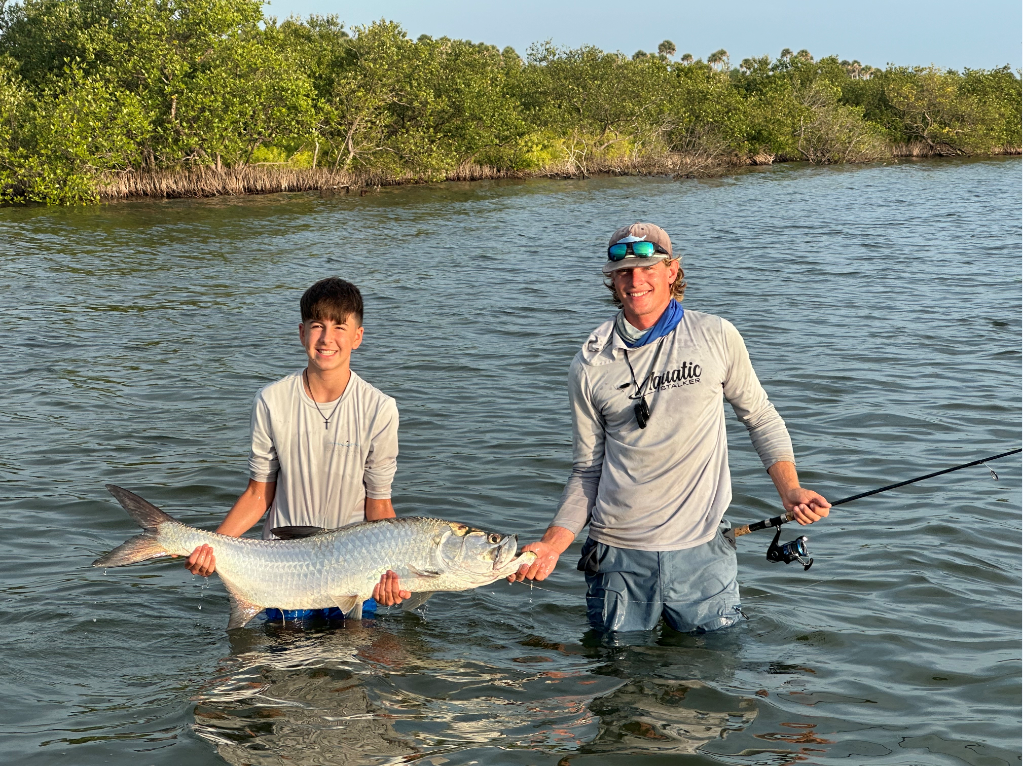 Man on a boat holding up a large fish he caught, wearing blue shorts and a pink cap, blue water in background.