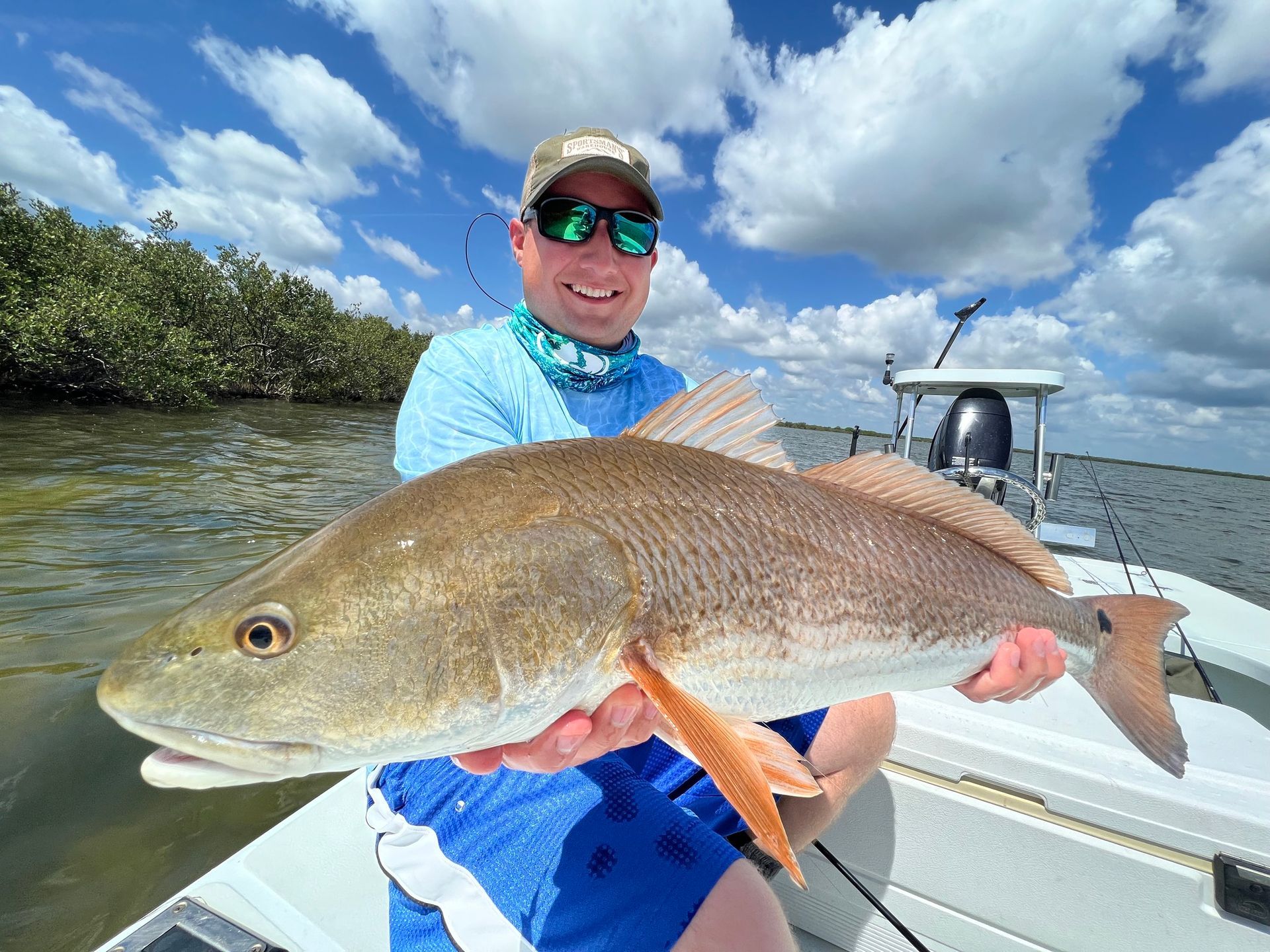 Man on boat smiling, holding a reddish-brown fish. Blue sky, water, and greenery in background.