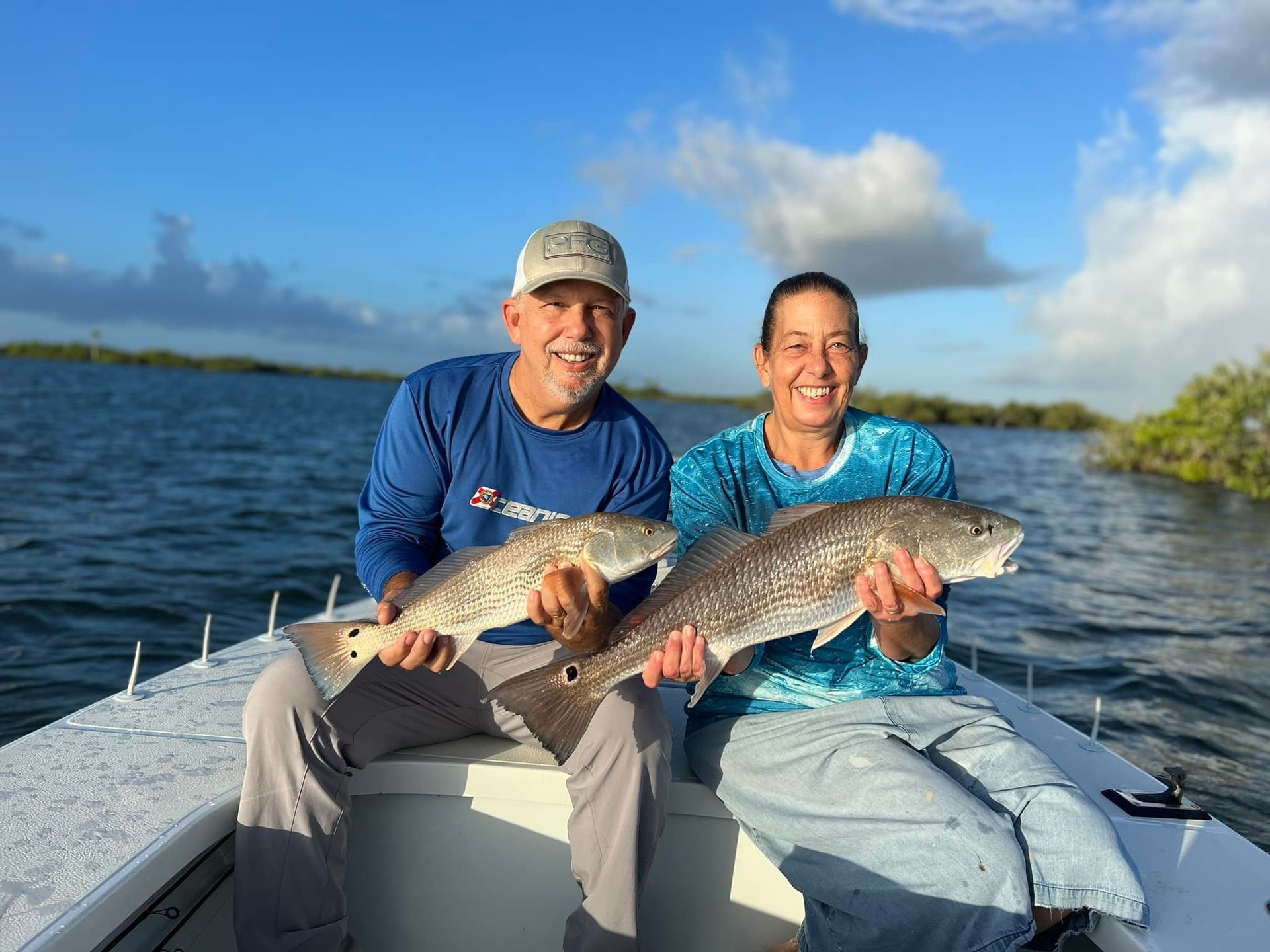 Two people on a boat display redfish they caught; blue water, partly cloudy sky.