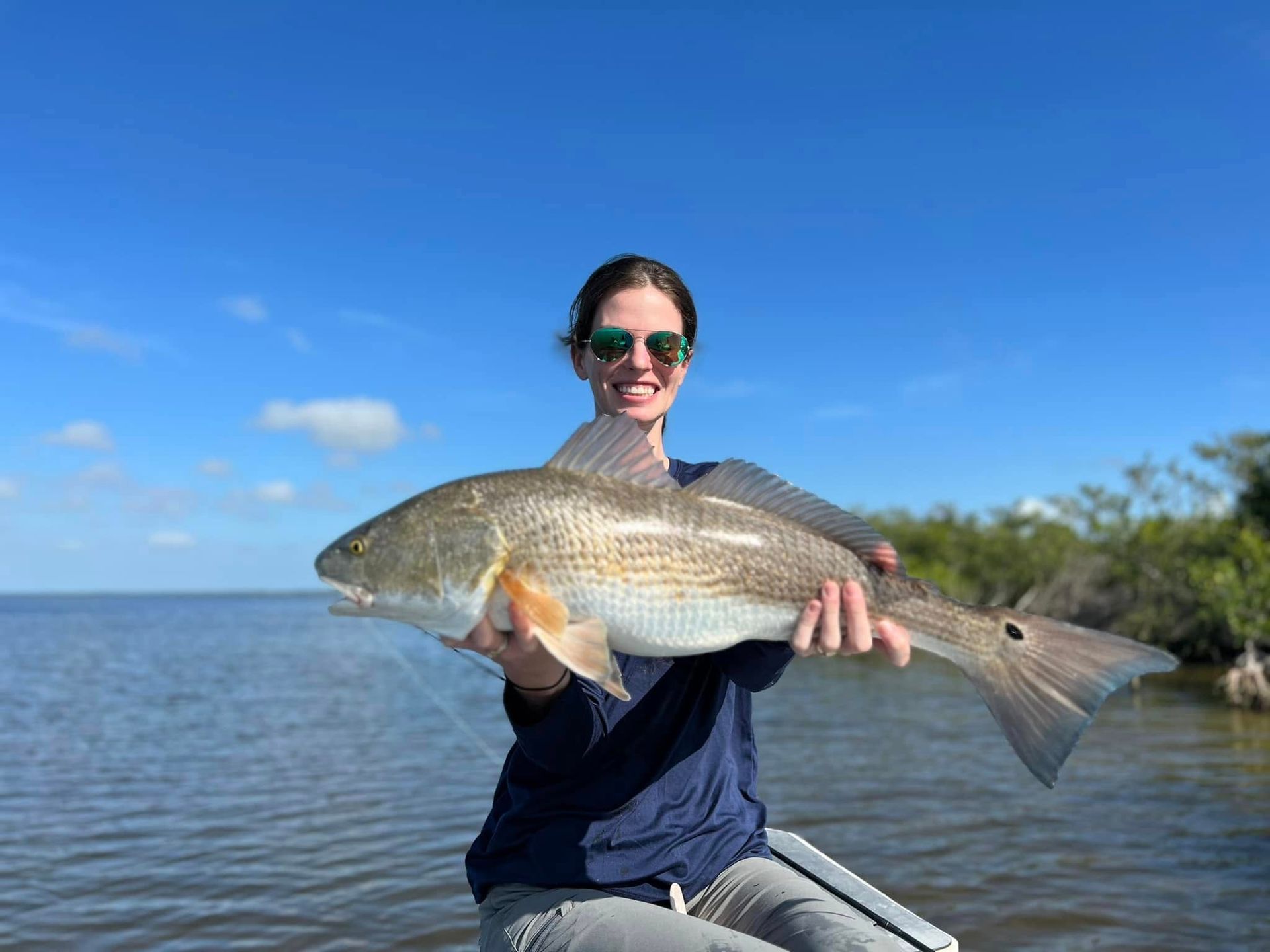 Woman in sunglasses smiles, holding a large redfish in a boat, near water and trees.