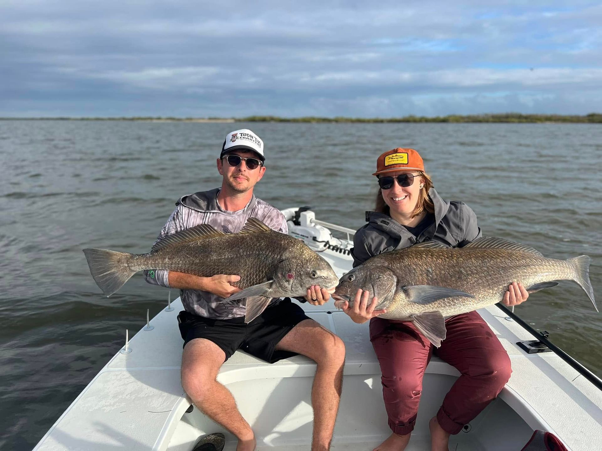Two people on a boat hold up large dark fish; river setting, overcast sky.
