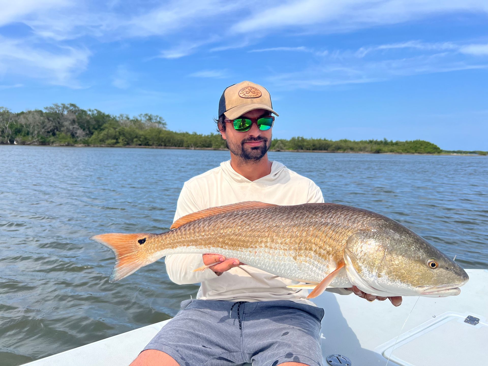 Man on boat holds up a large redfish with a neutral expression, cloudy day.